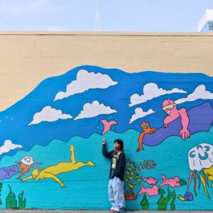 Sofia Angeles @s16topia pointing to her mural clouds, jellyfish an otter and a person in a canoe can be seen with Sophia pointing to the clouds.