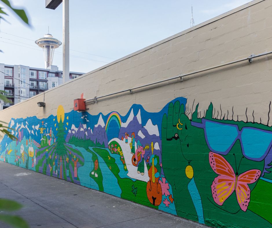Sofia Angeles mural on 113 Dexter Ave N that features a green character with their hand up to their ear while wearing glasses with sea creatures in bright contrasting colors.