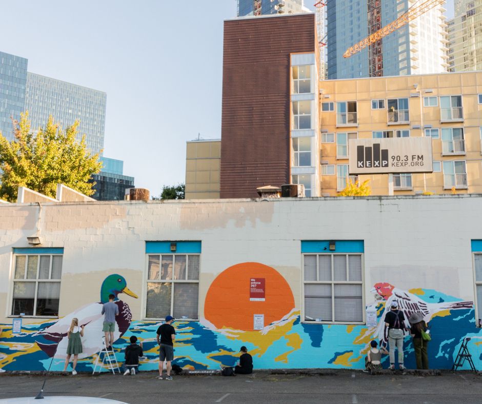 Sophia Huang's mural at 113 Dexter Ave N with an orange orb that resembles the sun in the middle and bright blue colors surrounding, with a mallard duck and a heron.