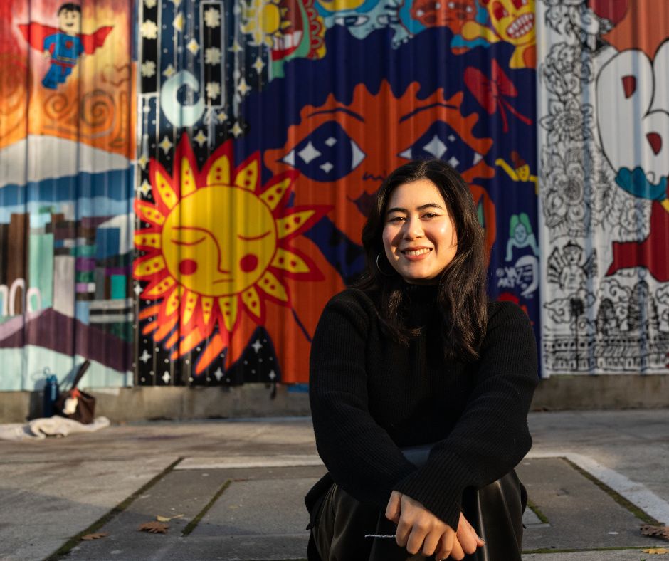 Yaz (aka Yasmin) sitting in front of her mural titled ‘Miss You, Sun’… Yaz says that in Seattle we have a lot of gloomy days and some people get seasonal depression, and I really wanted people to feel a sense of energy from my piece. So, it's a lady holding the sun and she's thinking, ‘I miss you sun’.”