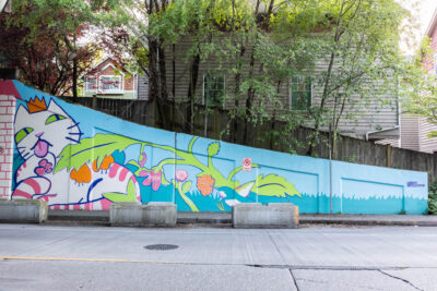 This image shows a colorful mural painted along a long retaining wall beside a residential street. The mural features a whimsical white cat wearing a gold crown, outlined in bold blue lines, stretching across the wall through a landscape of oversized leaves, flowers, berries, and flowing plant forms. The artwork uses a bright palette of turquoise, lime green, pink, orange, and blue against a light blue background, creating a playful and imaginative scene. The crowned cat appears to emerge from the foliage, symbolizing the neighborhood cats that inspired the design. Trees and houses rise above the wall, framing the mural within a lush urban environment. Concrete planters line the street in front of the artwork, while the mural's colors brighten the space into a welcoming and joyful public art experience.